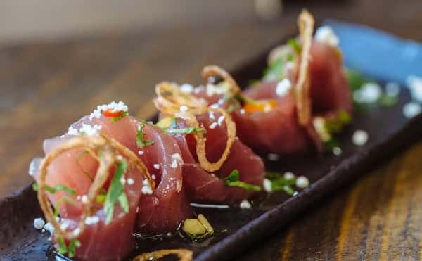 Close-up of fresh tuna sashimi rolls garnished with crispy shallots, herbs, and sea salt, served in a light soy-based dressing on a sleek black ceramic plate.