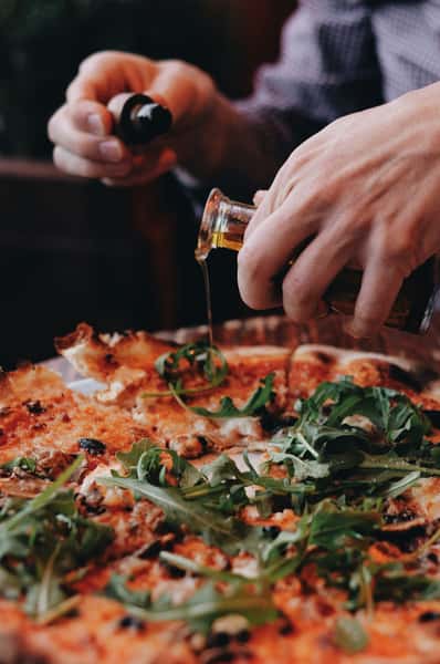 A hand pouring oil over a pizza topped with arugula