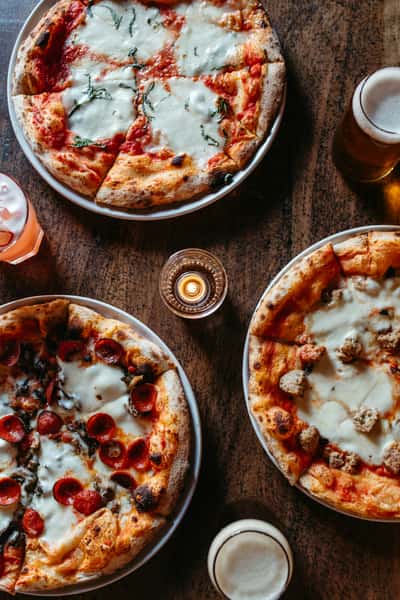 Three pizzas with various toppings accompanied by glasses of beer on wooden table