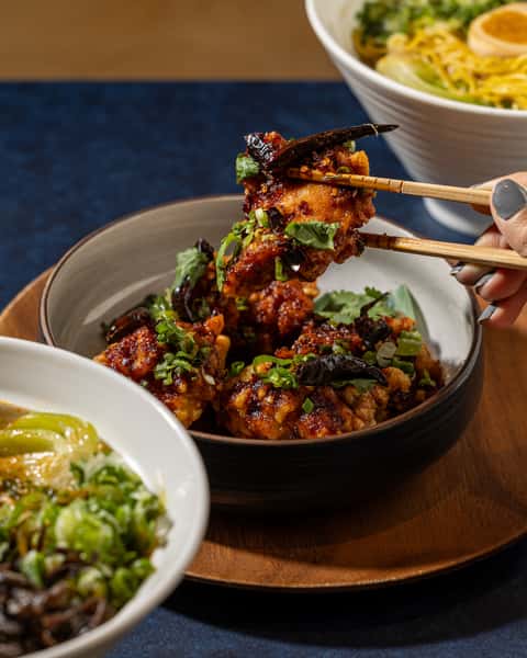 Spicy glazed karaage chicken garnished with herbs and dried chilies, lifted with chopsticks at a Japanese restaurant table.
