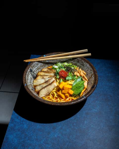 Japanese ramen bowl with grilled pork, wheat noodles, herbs, chili threads, and mixed toppings, styled on a deep blue table.