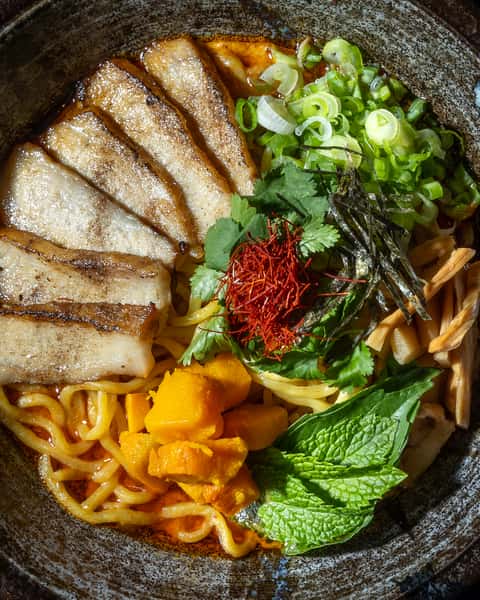Close-up of Japanese ramen with grilled pork belly slices, yellow noodles in a rich chili broth, topped with scallions, cilantro, bamboo shoots, seaweed, chili threads, and fresh herbs, served in a rustic ceramic bowl at a modern ramen restaurant.