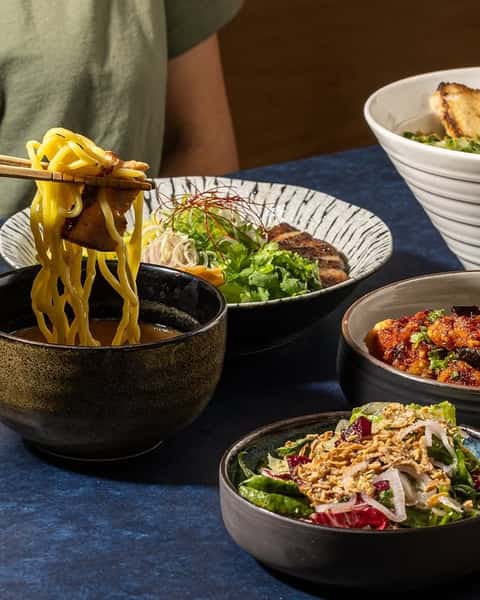Japanese dipping ramen noodles lifted with chopsticks from a rich broth, surrounded by small plates of glazed chicken, fresh salad, and ramen toppings on a shared restaurant table.