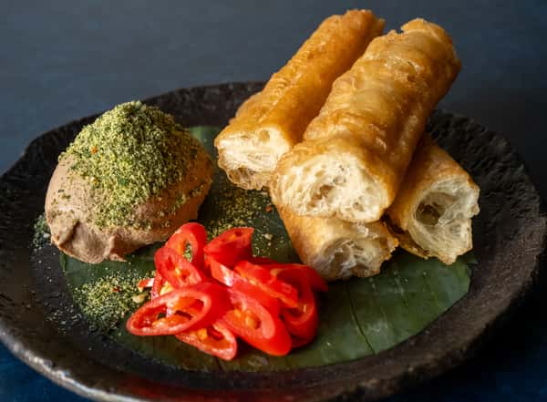 Japanese appetizer with airy fried bread sticks, seasoned spread, and pickled red peppers on a banana leaf.
