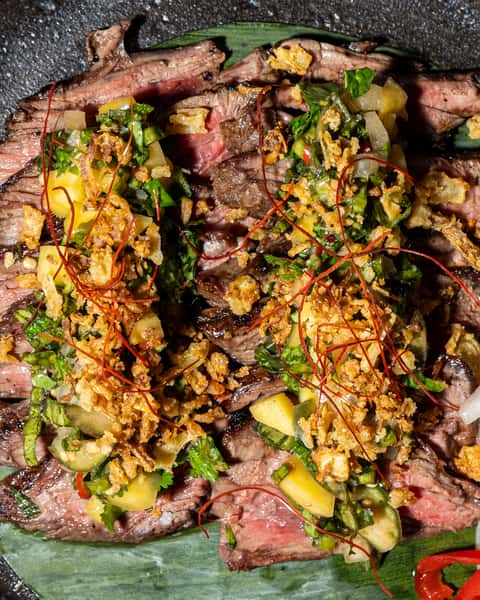 Sliced medium-rare steak topped with herbs, fried shallots, chili threads, and citrus relish, served on a ceramic plate.