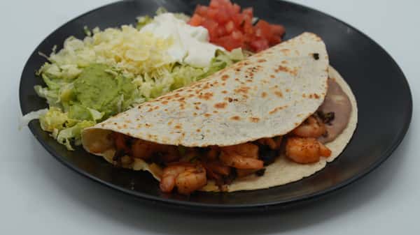 Grilled shrimp taco served on a lightly toasted flour tortilla with refried beans, accompanied by shredded lettuce, guacamole, sour cream, shredded cheese, and diced tomatoes—presented on a black plate for a flavorful and colorful Mexican dish.