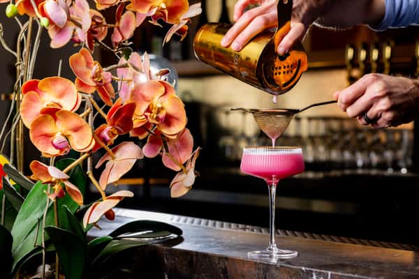 Bartender strains a pink cocktail into a coupe glass beside blooming orchids on a bar counter.