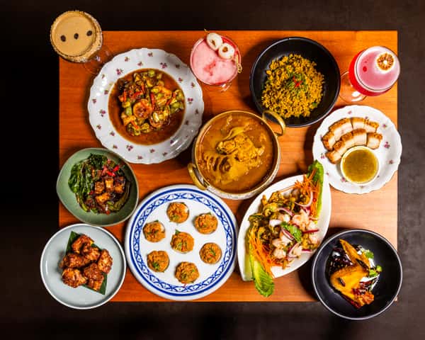 Overhead spread of Thai dishes and cocktails on a wooden table, featuring curries, salads, fried bites, and colorful drinks.