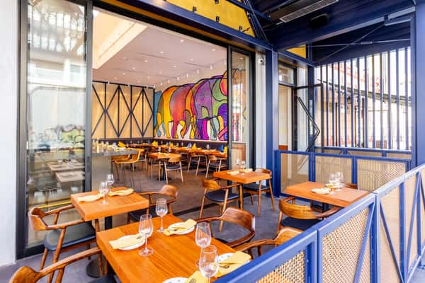 Outdoor restaurant patio with wooden tables and chairs, set for dining, looking into a modern interior with colorful elephant mural.