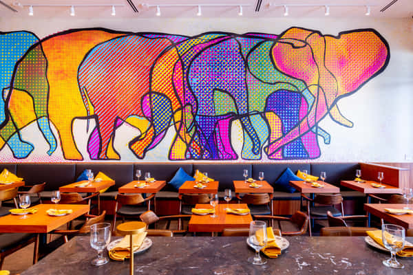 Modern restaurant dining room with wooden tables set with glassware and yellow napkins, beneath a vibrant, multicolored elephant mural spanning the wall.