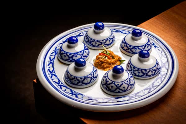 Blue-and-white ceramic platter with six covered dishes arranged around a central dollop of spicy relish on a wooden table.