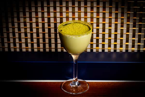Pale green cocktail in a coupe glass topped with matcha powder, set against a patterned bar backdrop
