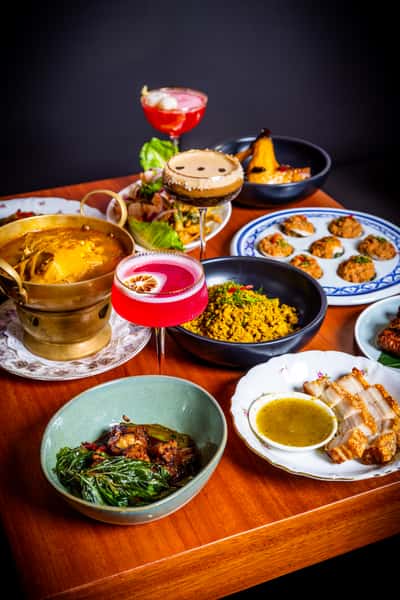 Colorful table spread with Thai-style dishes, curries, sliced pork, spiced dumplings, and vibrant cocktails arranged on a wooden table
