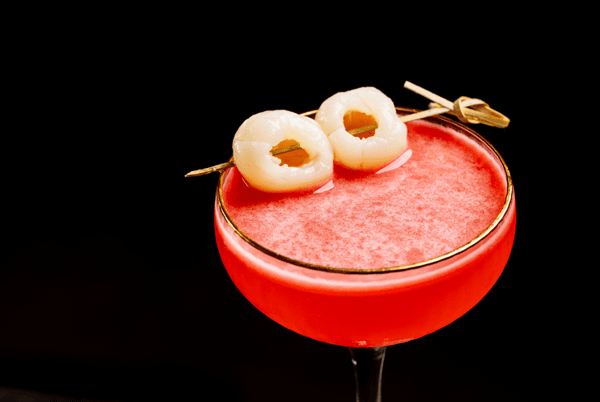 Red lychee cocktail in a coupe glass, topped with two lychees on a skewer, set against a dark background.