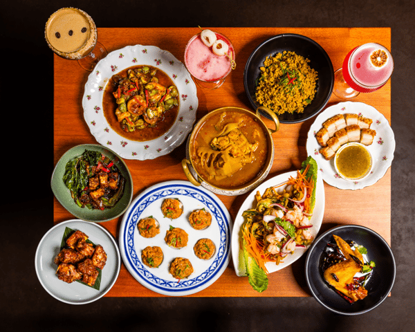 Overhead spread of Thai dishes and cocktails on a wooden table, featuring curries, salads, fried bites, and colorful drinks.