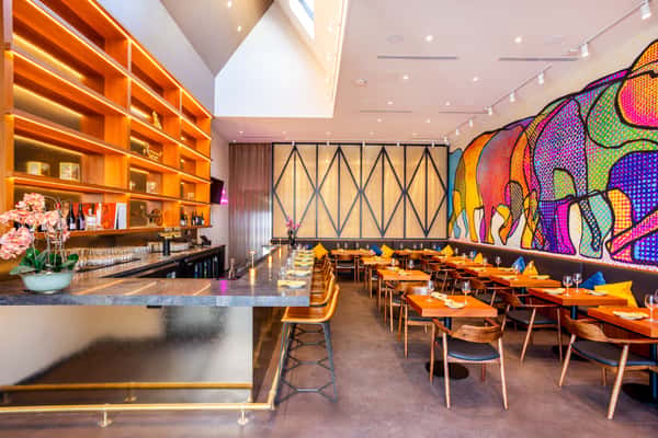 Modern restaurant interior with bar seating, wooden tables, warm lighting, and a colorful elephant mural along the wall