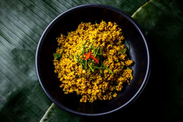Yellow minced curry garnished with herbs and red chili in a black bowl, set on a textured banana leaf background