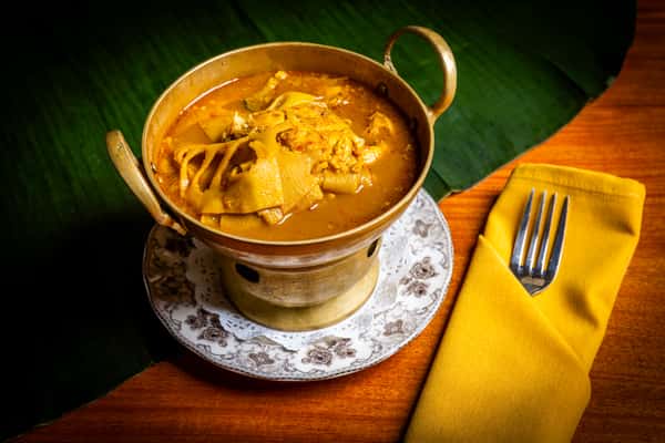 Brass pot of rich Thai curry with tender meat in golden broth on a patterned plate, set on a wooden table with banana leaf and yellow napkin