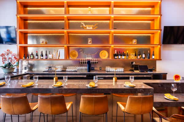 Warm modern bar with backlit wooden shelves, wine bottles, taps, and marble counter set with glasses and yellow napkins