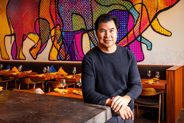 Man stands behind a restaurant counter with set tables and a colorful abstract mural featuring animal shapes in the background.