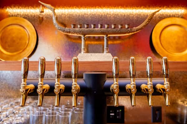 Row of polished brass beer taps above clean glassware, set against a warm, golden bar backdrop with decorative metal accents.
