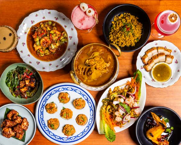 Overhead spread of Thai dishes and colorful cocktails on a wooden table with curries, seafood, pork, and small plates.
