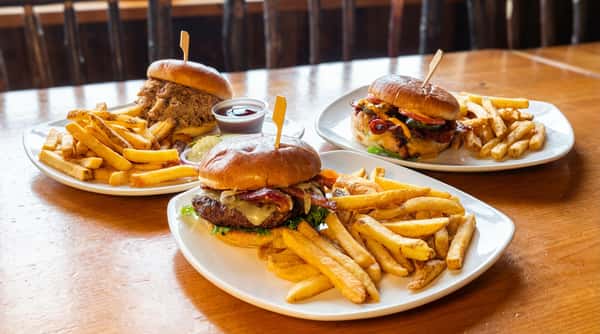 Three plates of burgers and fries on a wooden table. The front plate has a cheeseburger with bacon and lettuce.