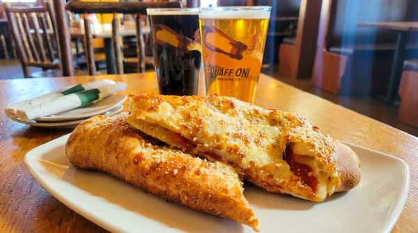 Two pepperoni and cheese stromboli on a white plate, served with two pints of beer, one light and one dark.