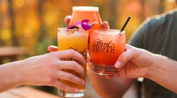 Three hands holding up different cocktails: an amber beer, a yellow drink with a purple orchid, and a reddish drink with a straw.