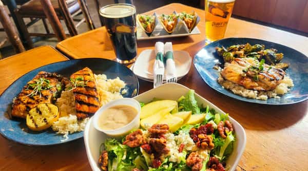 Tabletop full of food: grilled salmon with rice, chicken with brussels sprouts, tacos, salad with pears, blue cheese, walnuts, and cranberries, and two beers.