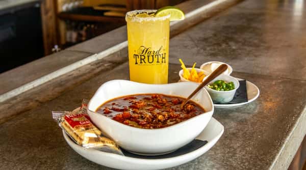 A bowl of dark red chili with a spoon, accompanied by side dishes of shredded cheese, sour cream, diced green peppers, and a margarita with a lime wedge.
