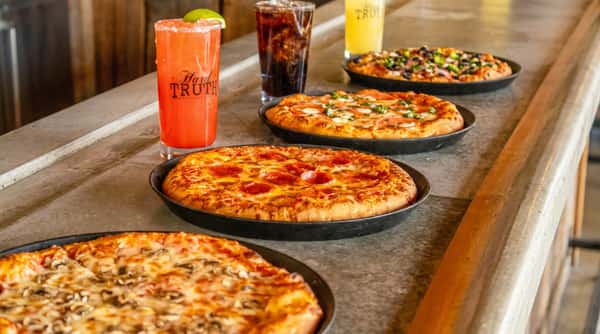 Four pizzas in cast iron pans, a red cocktail with a lime wedge, and two other drinks on a rustic bar.