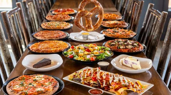 A long table filled with pizzas, a large pretzel, salad, nachos, and dessert slices.