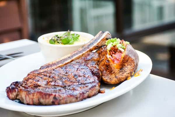 Wood-fired tomahawk steak with a fully loaded baked potato and creamy corn grits at Bovine's, a coastal cuisine favorite in Murrells Inlet, SC, known for its waterfront dining experience