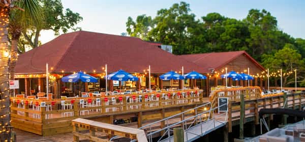 Evening view of Bovine's waterfront restaurant in Murrells Inlet, SC, glowing under string lights with blue umbrellas and dockside seating, perfect for wood-fired coastal cuisine by the inlet.