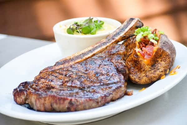 Juicy grilled tomahawk steak served with a loaded baked potato topped with cheese, bacon, and scallions, alongside creamy corn grits garnished with microgreens.