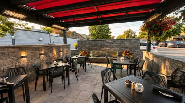 Outdoor restaurant patio with red canopy, dark tables and chairs, stone walls, and a glowing water feature.