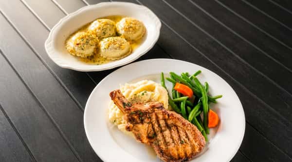 Grilled pork chop with mashed potatoes, green beans, and carrots, alongside ravioli in butter sauce.