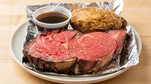 Slices of rare prime rib, a baked potato with salt, and a cup of au jus in a white takeout container lined with foil.