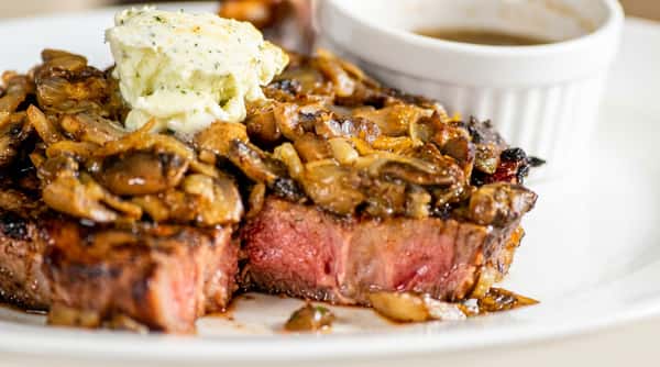 Medium-rare steak topped with sautéed mushrooms, onions, and melting herb butter, with a white ramekin.