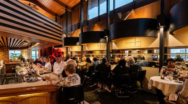 Darkly lit restaurant interior with patrons dining at tables and a long bar, featuring wooden ceiling accents and large black light fixtures.