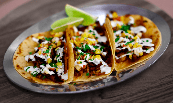Three street tacos filled with seasoned meat, corn salsa, fresh cilantro, and crema, served with lime wedges on a metal plate.