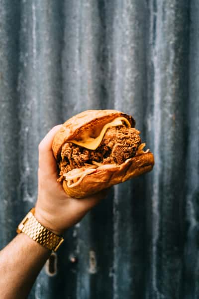 Hand holding a crispy fried chicken sandwich with melted cheese and onions against a corrugated metal background.