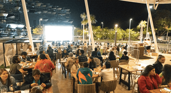 Crowded outdoor waterfront dining space at night with guests seated at tables and a large illuminated screen in view.