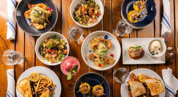 Overhead view of a wooden table filled with seafood pasta, fresh salads, sandwiches, dessert, and drinks arranged for a shared meal.