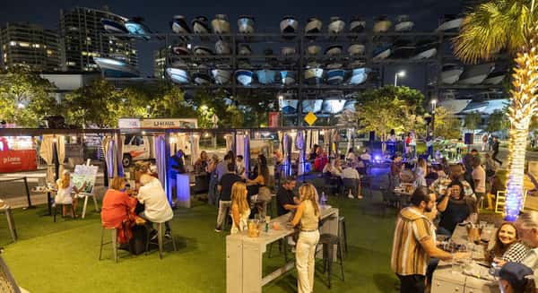 Evening outdoor event at a waterfront venue with guests socializing on a lawn, live music stage, palm trees, and stacked boats in the background.