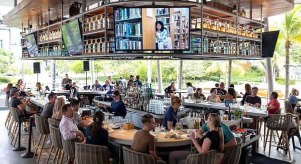 Open-air bar scene at a bayside restaurant with guests dining, large overhead TVs, and marina views through palm trees.