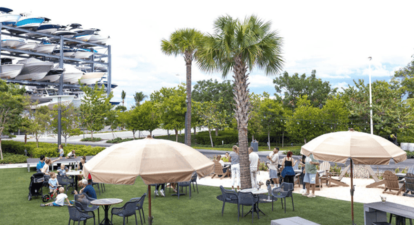 Outdoor waterfront dining area with palm trees, beige umbrellas, and families relaxing on a grassy patio near a marina.