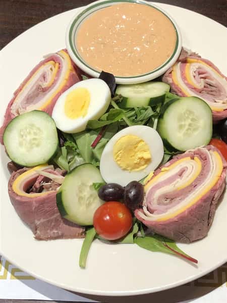 a chef's salad, with cucumbers, cherry tomatoes, and a hard boiled egg, with thousand island dressing on the side. Surrounding it is a ham, cheese and turkey roll that has been split into 4 pieces