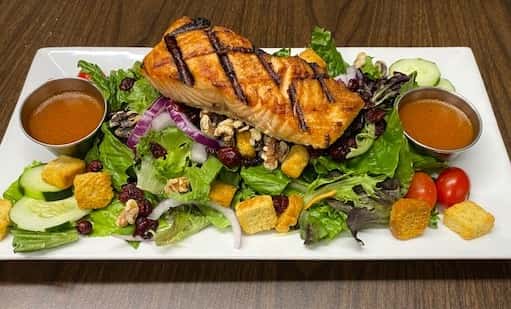 The image shows a grilled salmon fillet served over a fresh mixed salad with greens, sliced strawberries, mandarin oranges, and candied nuts. The salmon has visible grill marks and is plated in a rustic wooden bowl, giving it a fresh and hearty presentation.